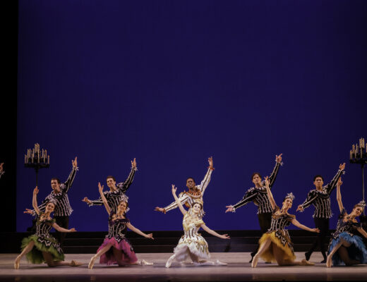 Eight male and eight female dancers pose in costume next to candelabras and a blue backdrop.