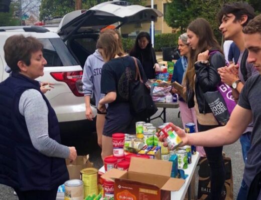 A group of people gather around a table of food at an outdoor food drive.